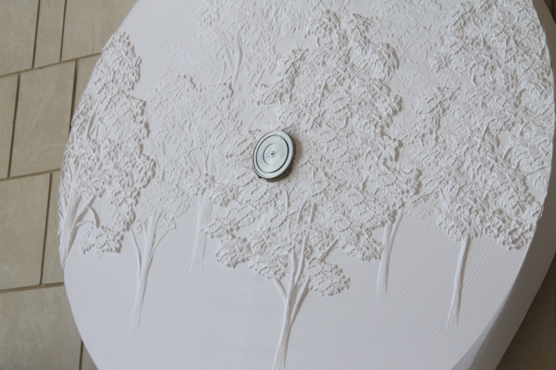 An antique typewriter set atop of a shelf that can be raised or lowered along a crankshaft attached to a sandstone wall. The typewriter is fed from a large roll of paper also attached to the wall. The surface of the paper roll features a low-bas relief carving of poplar trees.