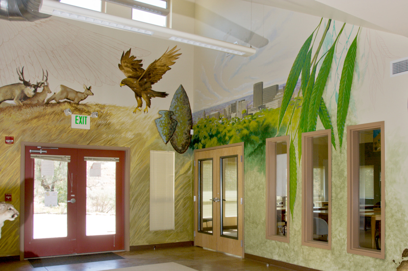 The main office of the Boise Foothills Learning Center. On the walls is painted a mural depicting a grassy hill with a heard of deer, a hawk, and a view of the Boise skyline. There are also overlying images of a flint arrowhead and spearhead, as well as a closeup of willow leaves.