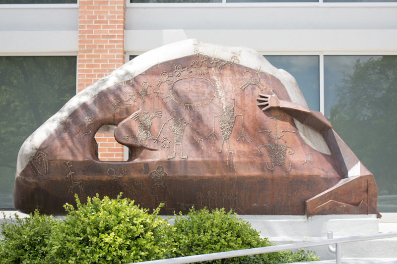 A weathered and stainless steel sculpture that depicts a Native American clinging to a rock formation that features petroglyphs. 