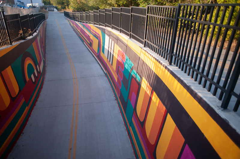 A mural on the retaining walls of a bike path heading into a tunnel. The design consists of lines and abstract shapes in the colors black, white, red, orange, yellow, teal, and magenta.