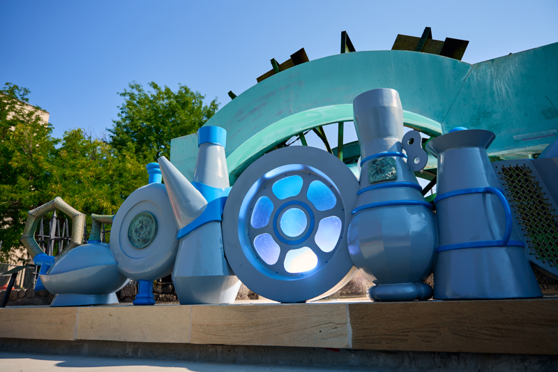 A sculpture depicting a lineup of 6 water clock vessels from different cultures in front of the waterwheel at Boise's C.W. Moore Park.