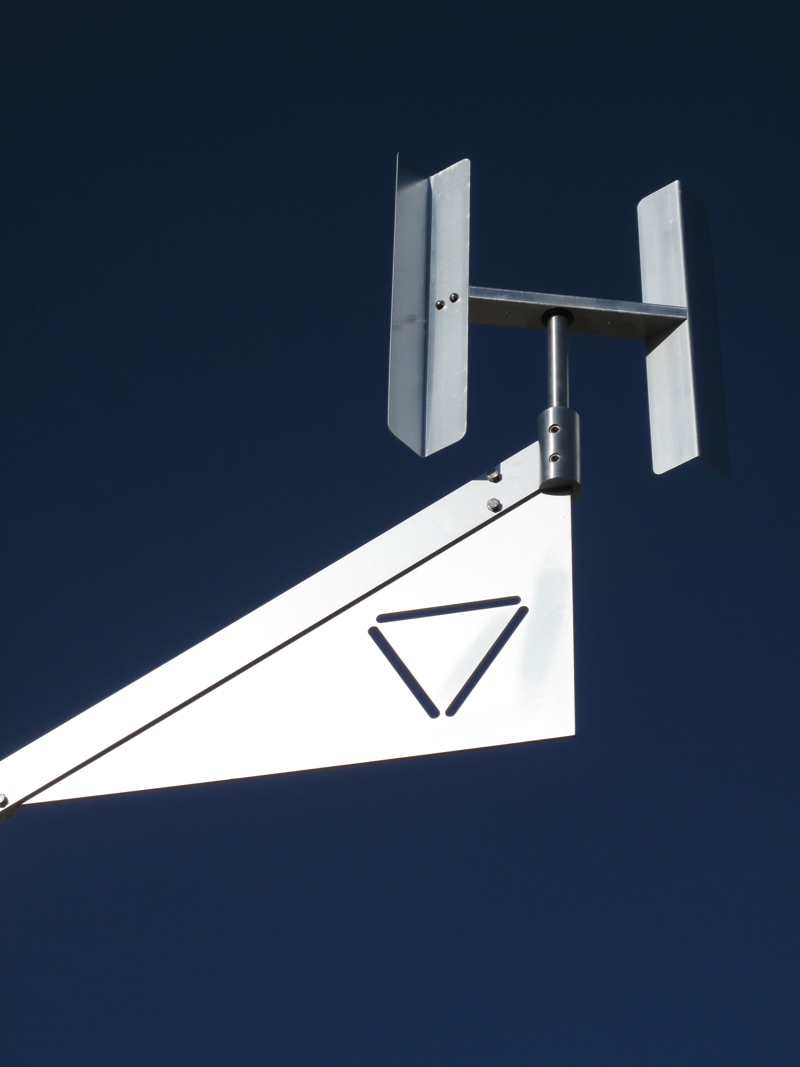 An aluminum kinetic sculpture representing a water molecule. At its central axis point is the letter O from which stems two arms with the letter H at their end. This images shows a detail closeup of the end of one of the arms which has a symbol of a triangle meant to symbolize the solid state of water.