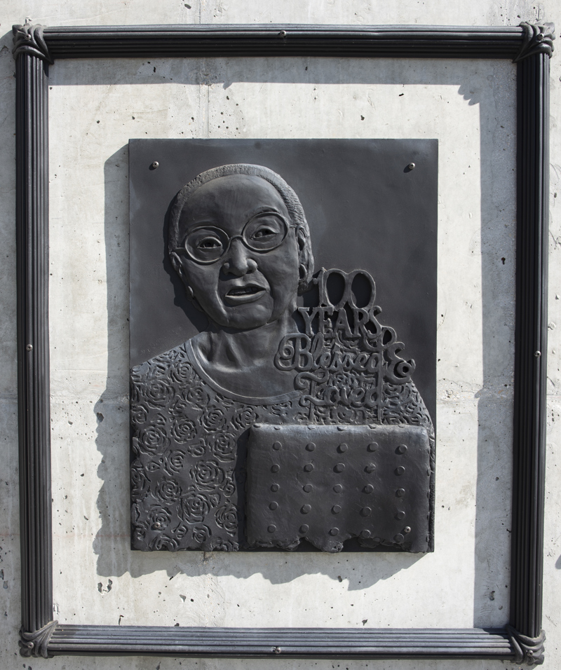 A low bas-relief bronze resin sculpture depicting a bust photo of an elderly woman next to a cake. On the cake is a topper which reads "100 Years Loved & Blessed." The sculpture is rectangular with a rough edge and is surrounded by a separate ornate frame.