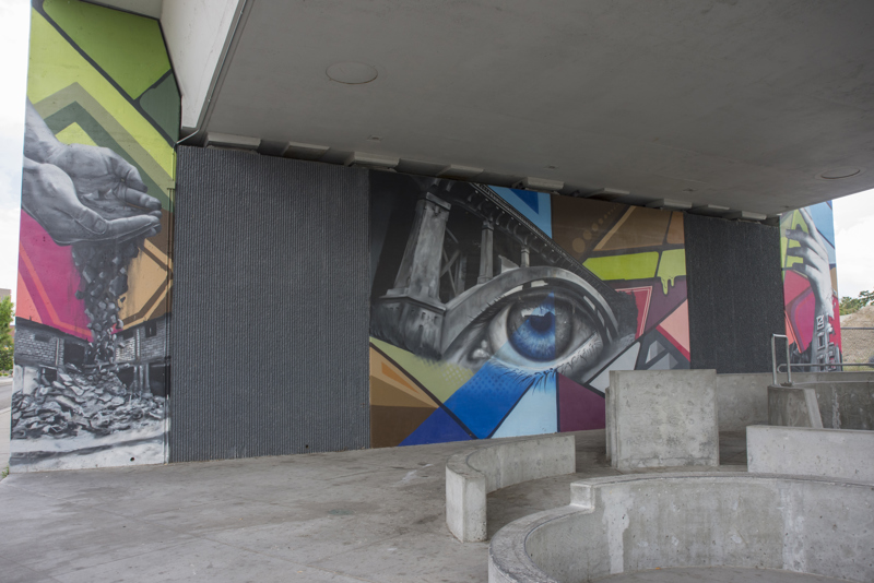 A mural painted in three parts on the wall of a highway overpass. On the left is the image of a pair of hands holding soil that slips through its fingers, turning into building rubble. In the middle is an image of a single eye situated beneath the arch of a bridge. On the right is an image of a hand and forearm turning into the crumbling facade of an abandoned building. 
