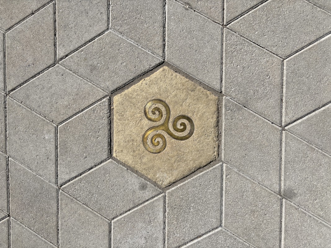 An image of a hexagonal sidewalk paver with a brass inlay of an Irish symbol.