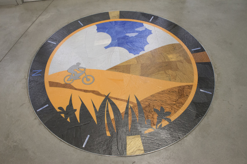 A floor inlay medallion depicting a biker in the Boise foothills. Around the outside of the medallion are the cardinal directions with the letter N indicating north. 