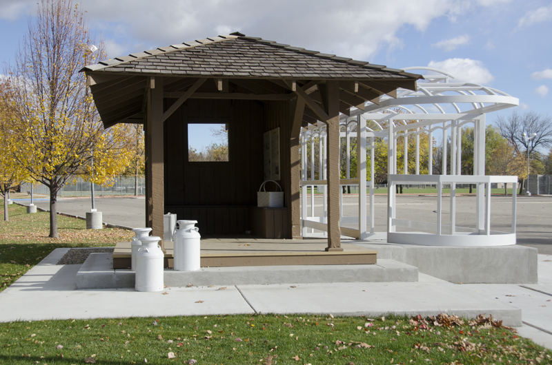 A historic streetcar station shelter made of wood. Outside are old metal milk containers painted white. In the interior are a suitcase and a basket made of metal painted white. Adjacent to the station is the metal skeletal frame of a streetcar painted white.