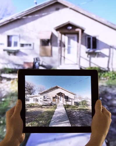 Hands holding a smart tablet displaying a photo of the James Castle House. Behind the tablet is the actual James Castle House.