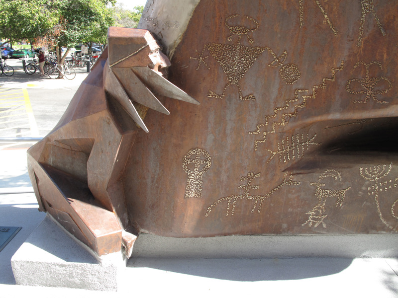A weathered and stainless steel sculpture that depicts a Native American clinging to a rock formation that features petroglyphs. 