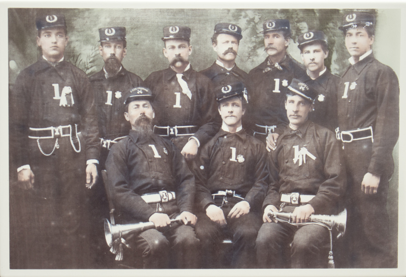An image of several firefighters in uniform sitting for a portrait in 1876.
