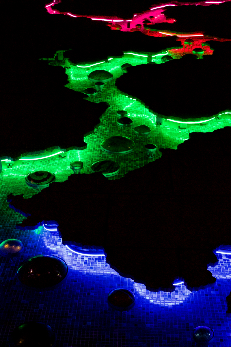 A large scale installation spanning nearly fifty feet of a building corner made up of rectangular granite slabs. Running up the center of the wall is a cutout shape of a large shape resembling a crack or a river and its tributaries. Recessed in this "crack" is a blue glass-tiled surface and circular, colorful blown glass components. Obscured beneath the cut edges of the granite, outlining the shape of the crack, are LED ribbons that illuminate the installation at night.