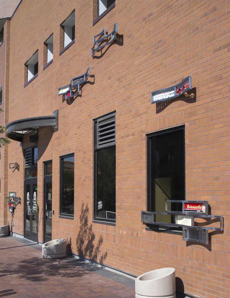 Six groupings of wavy stainless steel frames affixed to the exterior red brick wall of Boise City Hall. Some frames contain written and visual content with Chinese figures and historical references.