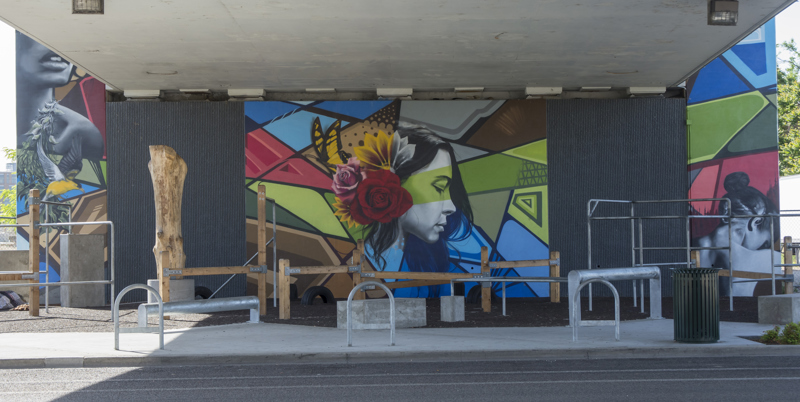 A mural painted in three parts on the wall of a highway overpass. On the left is the image of a yellow bird in flight, overlapping a leafy branch and a woman's bust. In the middle is an image of a woman in side profile with flowers in her hair. On the right is an image of a woman's upper back as she looks down. 