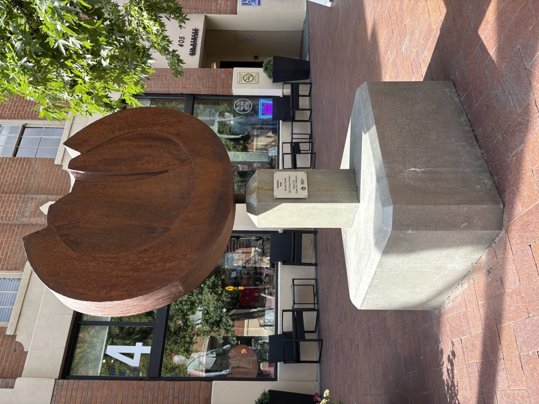 An abstract sculpture made of weathered steel attached to a rectangular column base made from sandstone.