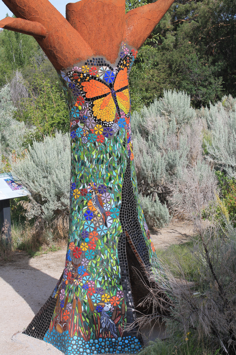 A life size sculpture of a dead standing hollow tree trunk. The sculpture is made of weathered steel and covered in glass tile mosaic depicting various lifeforms (i.e. birds, insects, and plants) that make use of dead or fallen trees in their habitat.