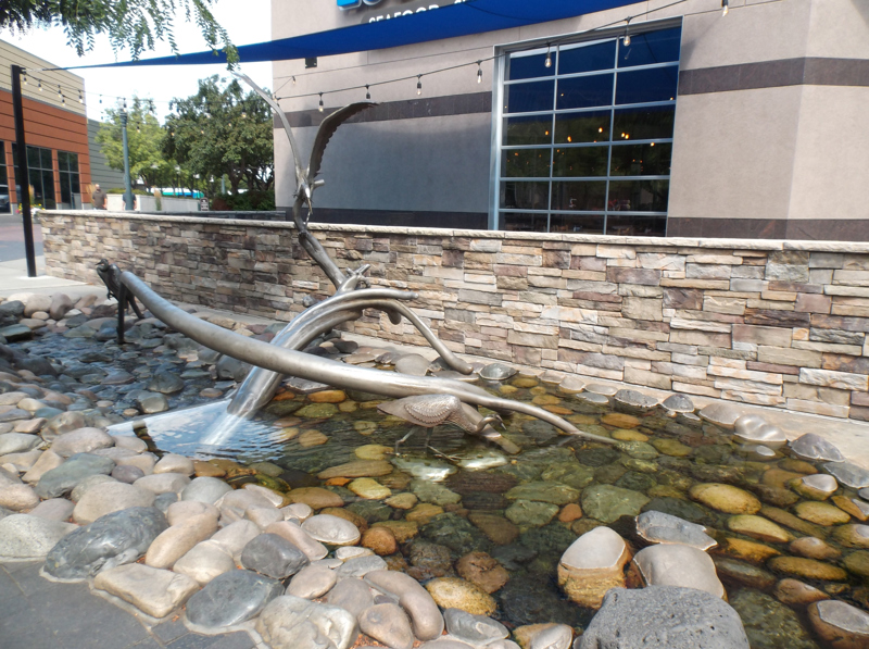 A water feature with an accompanying sculpture. Water flows as a small stream over smoothed river rocks of different colors beneath a stainless steel sculpture of three great blue herons on and around two fallen trees.