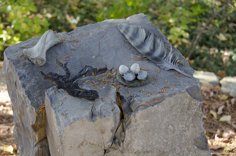 A basalt stone pedestal with four small patinated bronze-cast naturalistic sculptures attached to the top. The small sculptures are a feather, a nest of eggs, a charred branch, and a bone.