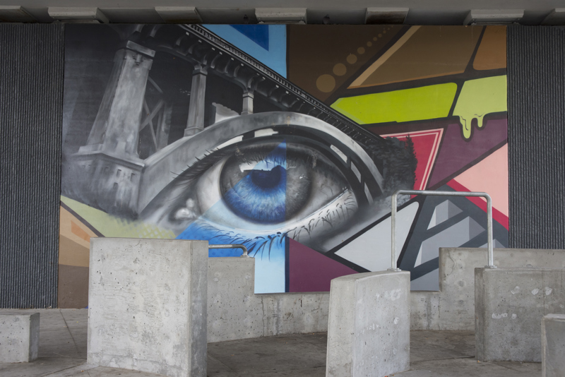 A mural painted in three parts on the wall of a highway overpass. On the left is the image of a pair of hands holding soil that slips through its fingers, turning into building rubble. In the middle is an image of a single eye situated beneath the arch of a bridge. On the right is an image of a hand and forearm turning into the crumbling facade of an abandoned building. 