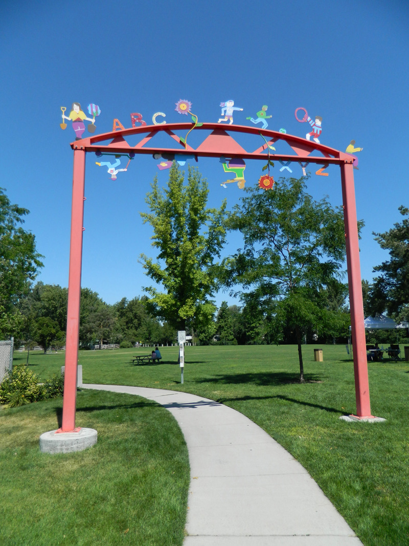 A standing metal arch over a park walkway. The arch is painted red with various colorful figures of children at play attached to the top.