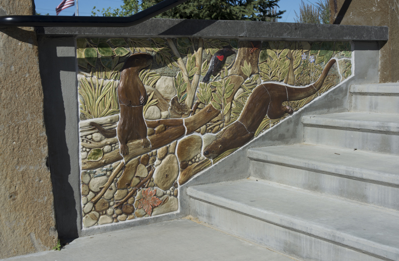 An inner wall of an exterior stairwell made with a sandstone top and a base decorated with painted low bas-relief ceramic sculptures depicting two river otters and a red-winged blackbird on the edge of a river. 