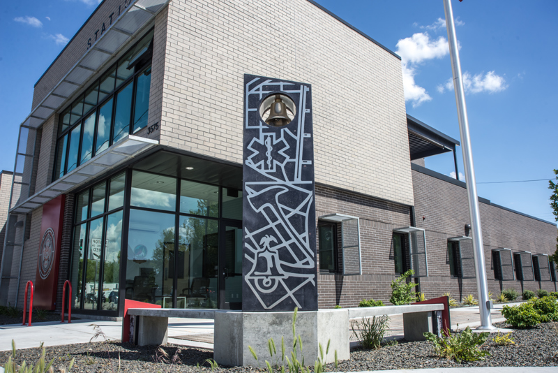 A corner bend made of cement, composite panels with printed images, and stainless steel cutouts of symbols and maps relating to Boise's first responders, namely firefighters. At the corner of the bend is a standing panel with a brass bell at its top. 
