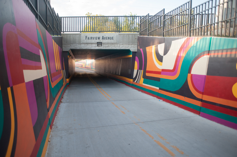 A mural on the retaining walls of a bike path heading into a tunnel. The design consists of lines and abstract shapes in the colors black, white, red, orange, yellow, teal, and magenta.